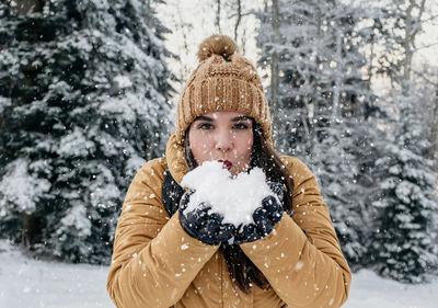 Portrait of beautiful young woman standing outdoor in snowy meadow in woods, blowing snow kiss