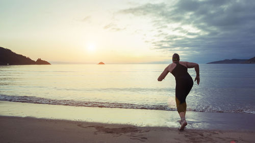 Back view of unrecognizable professional swimmer in swimsuit and cap ready for running in sea water during training at seaside at sunset