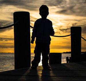 Silhouette boy standing on jetty in sea against orange sky during sunset