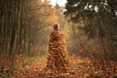 Young woman covered with leaves standing at forest during autumn