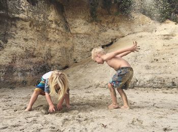 Siblings playing on sandy beach against rock formation