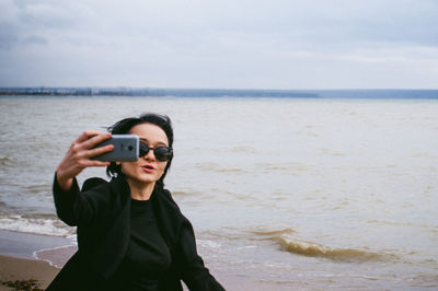 Woman taking selfie through mobile phone at beach against sky