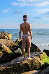 Rear view of shirtless man standing on rocky shore against blue sky