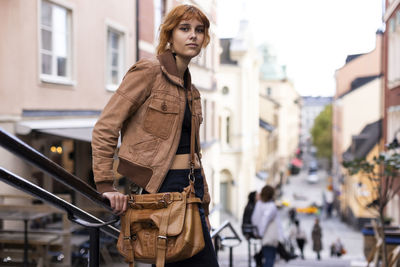 Portrait of young woman with bag and jacket standing near railing in city