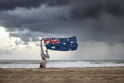 Woman holding flag while exercising on beach against sky