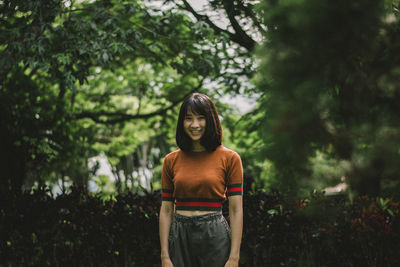 Portrait of smiling young woman standing against trees in park