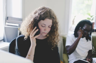 Young businesswoman talking on mobile phone with colleague in background at creative office