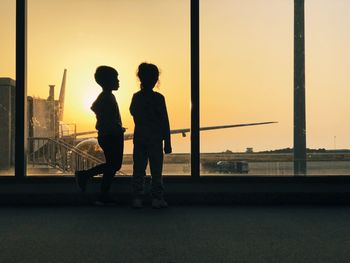 Boy and girl against window at airport during sunset
