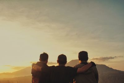 Rear view of friends with arms around standing against sky during sunrise