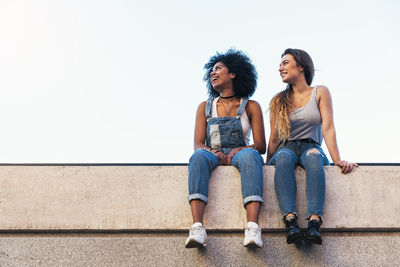 Full length of friends sitting on retaining wall against clear sky