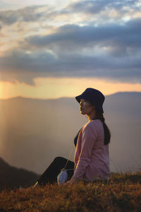 Side view of woman siting on grass against sky during sunset