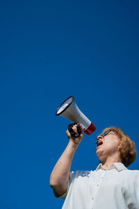 Low angle view of young man using mobile phone against clear blue sky