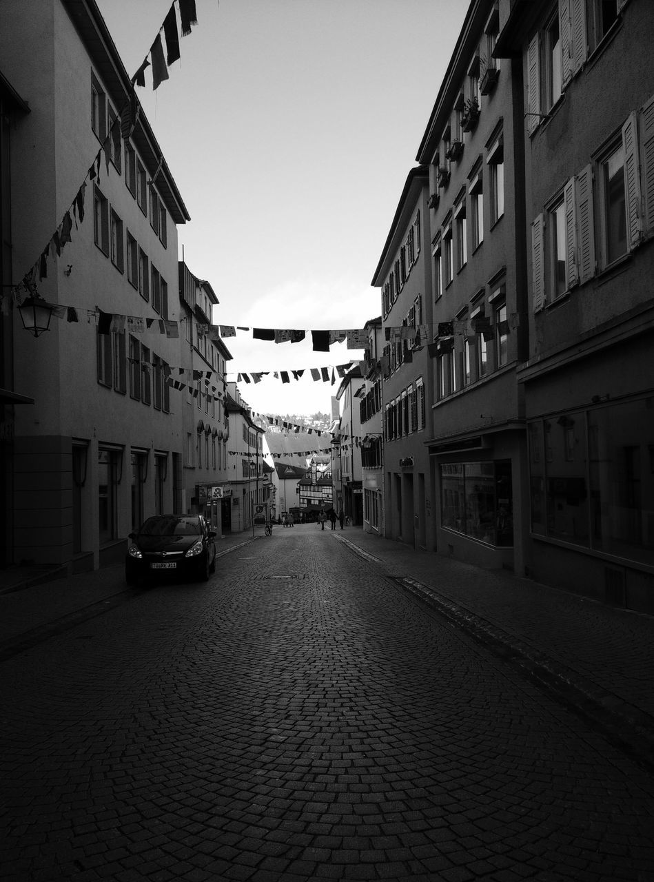 building exterior, architecture, built structure, street, the way forward, clear sky, city, transportation, building, residential structure, diminishing perspective, residential building, car, land vehicle, road, incidental people, sky, mode of transport, vanishing point, cobblestone