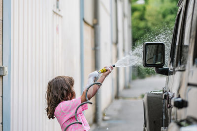 Side view of cute girl with hose and soap on road while washing the car