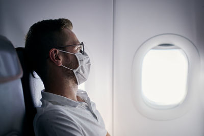 Passenger with protective face mask. young man man looking out window of airplane. 