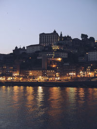 Illuminated buildings by river against sky at dusk