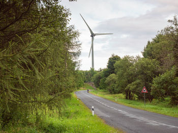 Asphalt road between trees up to hills with wind turbines on the peaks