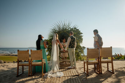 People sitting on deck chairs by sea against clear sky