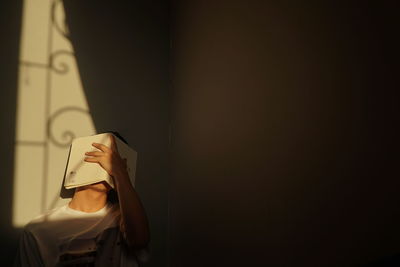 Man covering face by book while sitting by wall at home