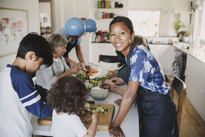 Portrait of happy woman standing with family preparing food at table in kitchen