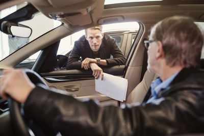 Salesman looking at senior man sitting in car at dealership shop