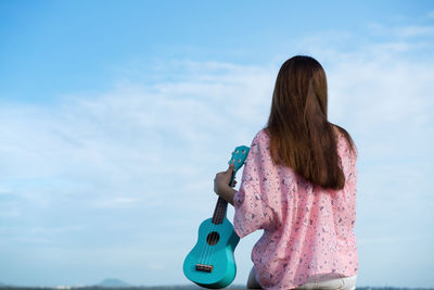 Rear view of woman holding ukulele while sitting against sky