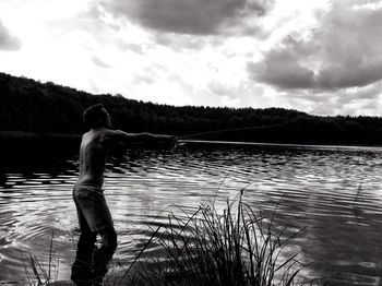 Shirtless young man fishing in lake against sky