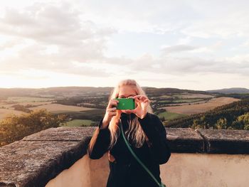 Young woman photographing on mobile phone against sky