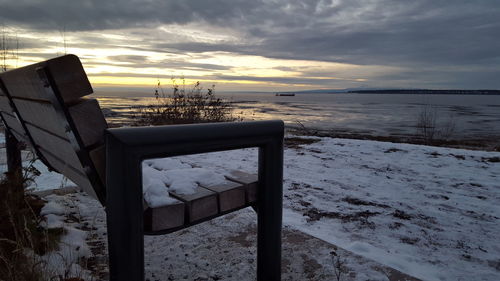 Empty bench at beach against cloudy sky during winter