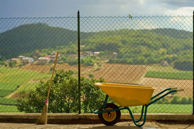 Wheelbarrow and broom by fence against landscape