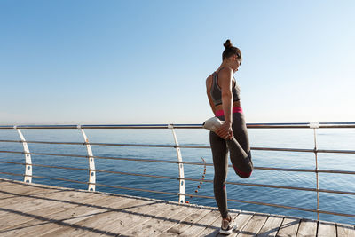 Rear view of woman exercising by railing while standing against sea