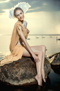 Portrait of young woman sitting on rock in sea against sky