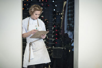 Female restaurant owner using smart phone standing with clipboard on doorway