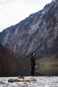 Side view of male tourist enjoying amazing landscape of calm lake and majestic mountains with forested slopes and snowy peaks in cloudy autumn day