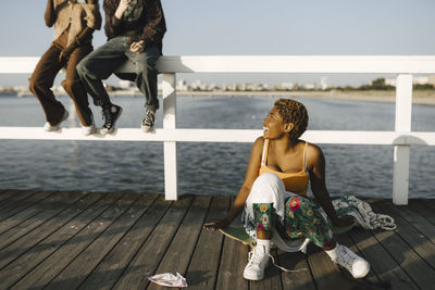 Smiling non-binary woman sitting on skateboard while looking at friends