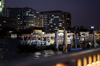 Boats moored in river against illuminated buildings in city at night