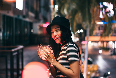 Portrait of smiling young woman holding illuminated string light while standing in city