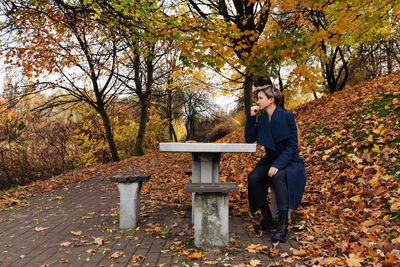 A short-haired woman in a coat sits at a table in the park in the fall