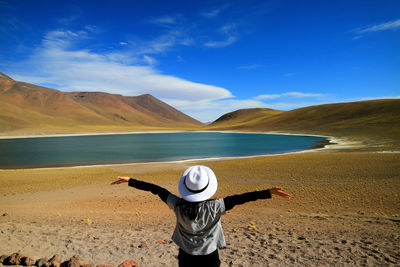 Rear view of mature woman with arms outstretched standing near lake against blue sky