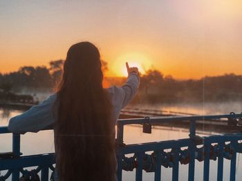 Rear view of woman standing by railing against sky during sunset