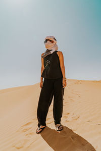 Full length of women wearing sunglasses with black dress and white turban standing on sand dune