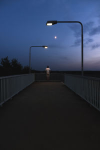 Rear view of man standing on bridge amidst illuminated street light against sky at dusk