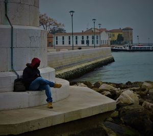 Man sitting on retaining wall by sea against sky in city