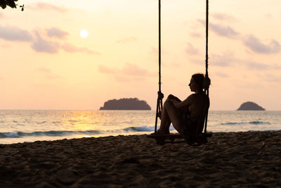 People sitting on shore at beach against sky during sunset