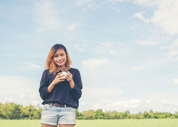 Smiling young woman with coffee cup standing against sky