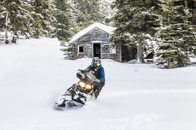 Confident man on snowmobile with old cabin behind.