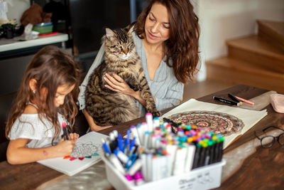 Daughter drawing on paper while mother holding cat at home
