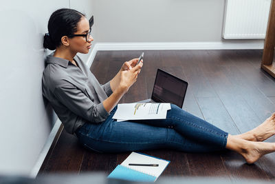 Businesswoman using mobile phone while sitting with documents and laptop on floor at home