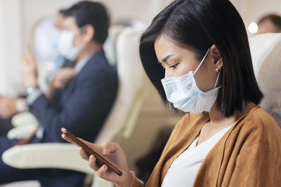 Passenger wearing face mask using mobile phone on airplane during covid pandemic to prevent 