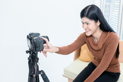 Smiling young woman holding camera while standing against white wall